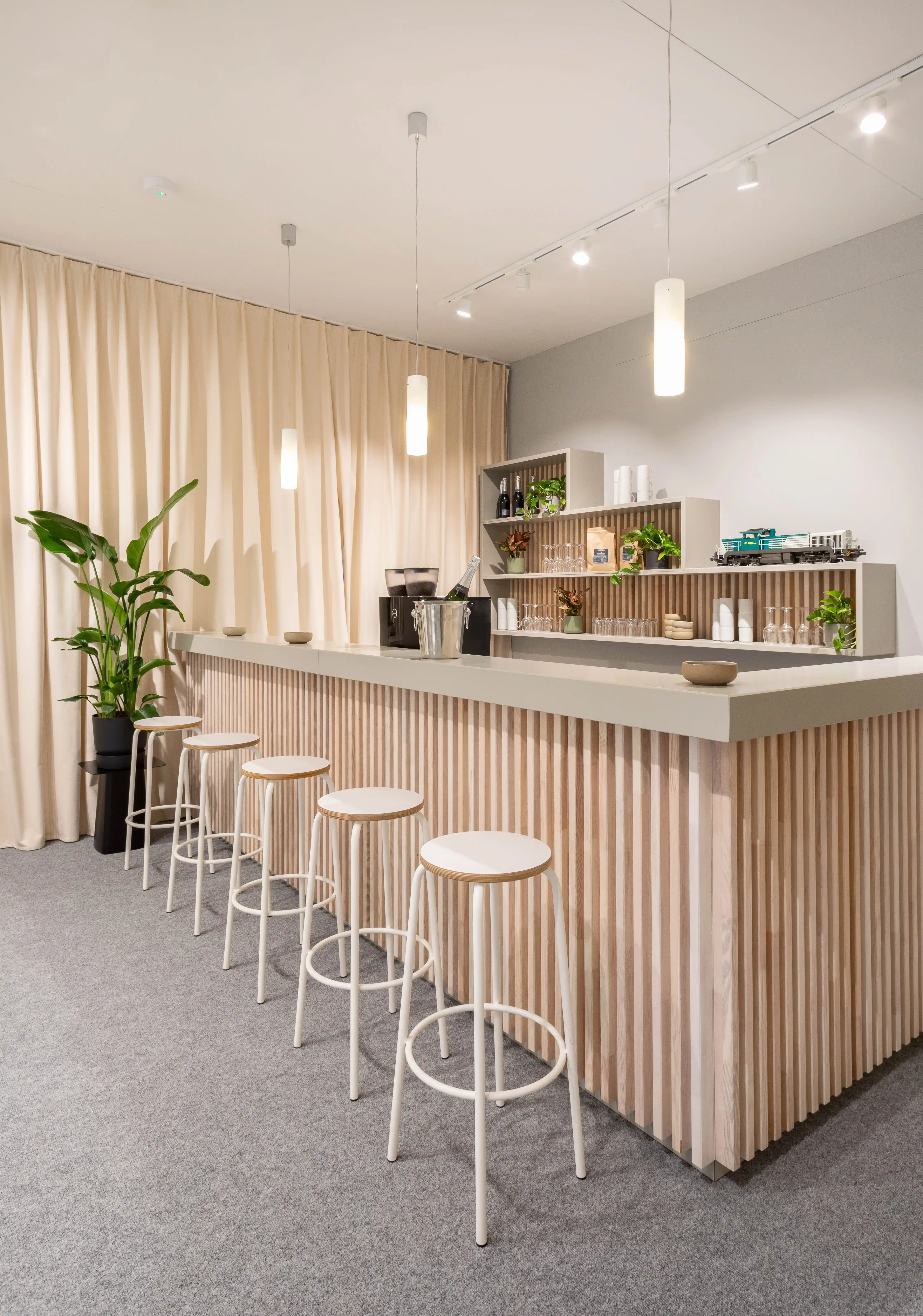 Bright bar with wood-slat front, white bar stools and plants in front of cream curtains in the Vossloh house.