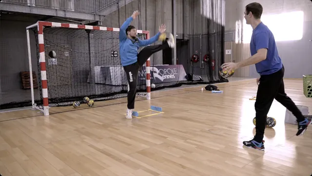 A handball goalkeeper raises a leg and both hands to block a ball during a close-range training drill with a coach in front of the goal.