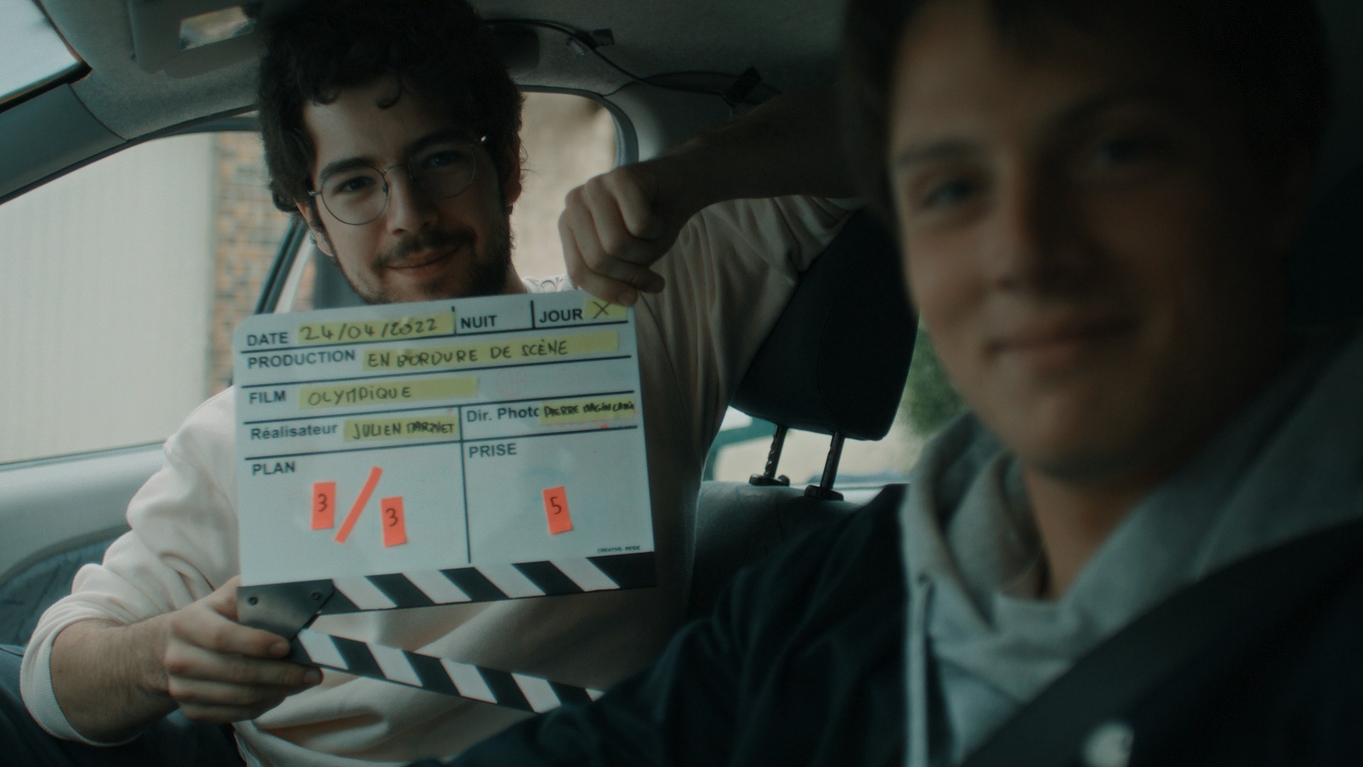 Scène de tournage à l'intérieur d'une voiture pour le film 'Olympique', membre de production tenant un clap de cinéma.