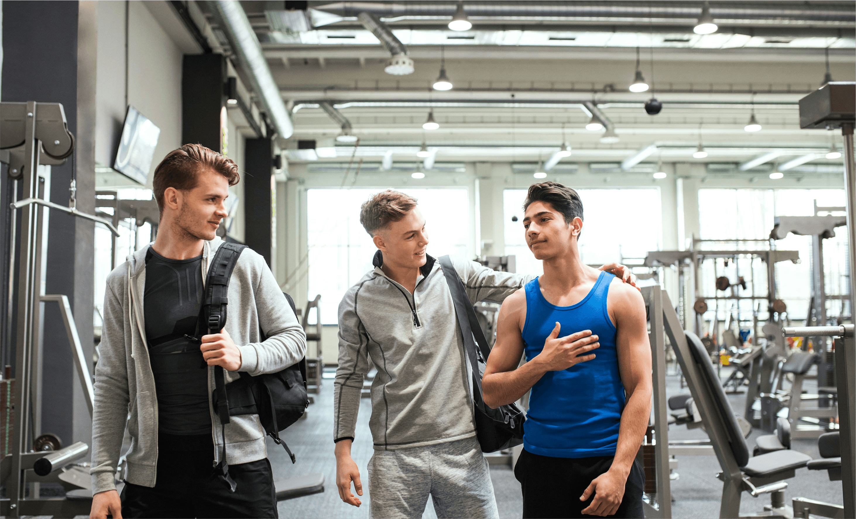 Three men talking and smiling in a modern gym, discussing workout plans with their trainer.