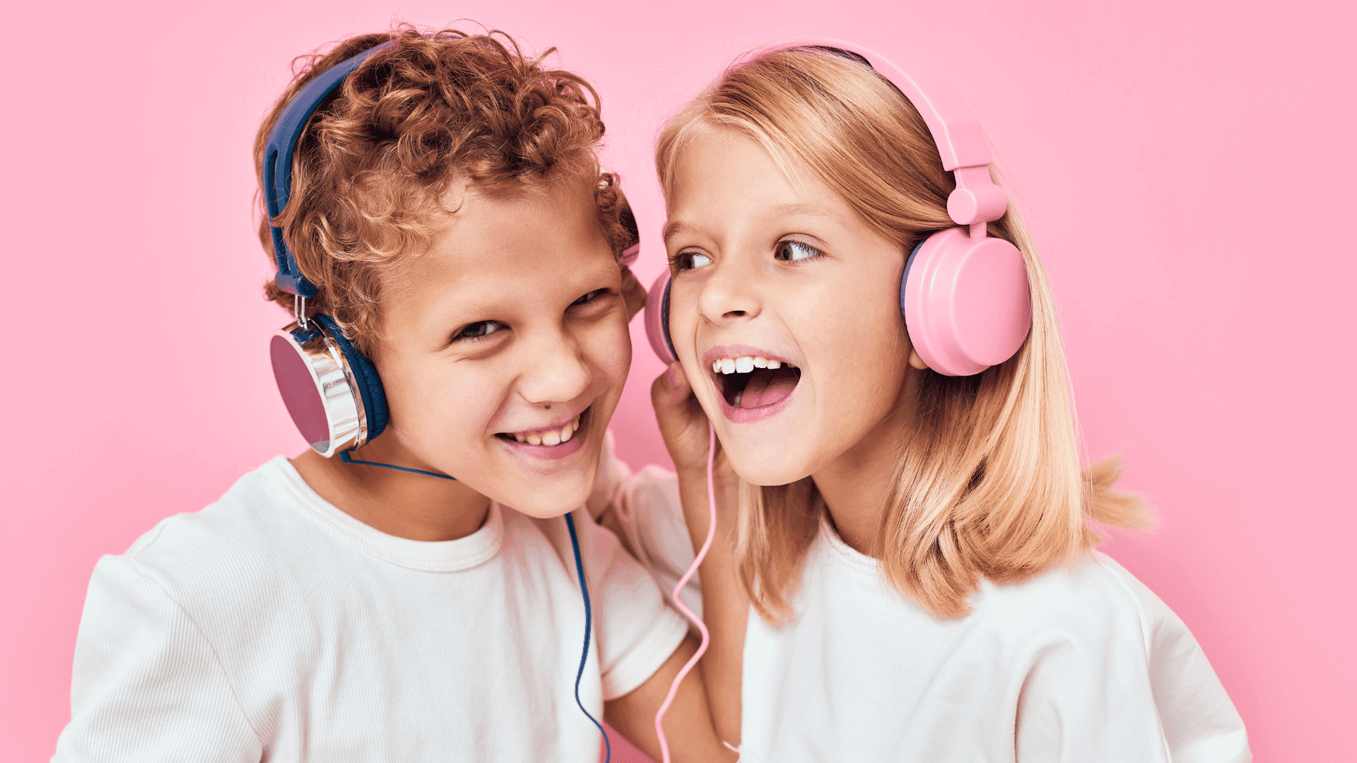 Two children standing in front of a pink background, wearing headphones, both smiling and looking happy.