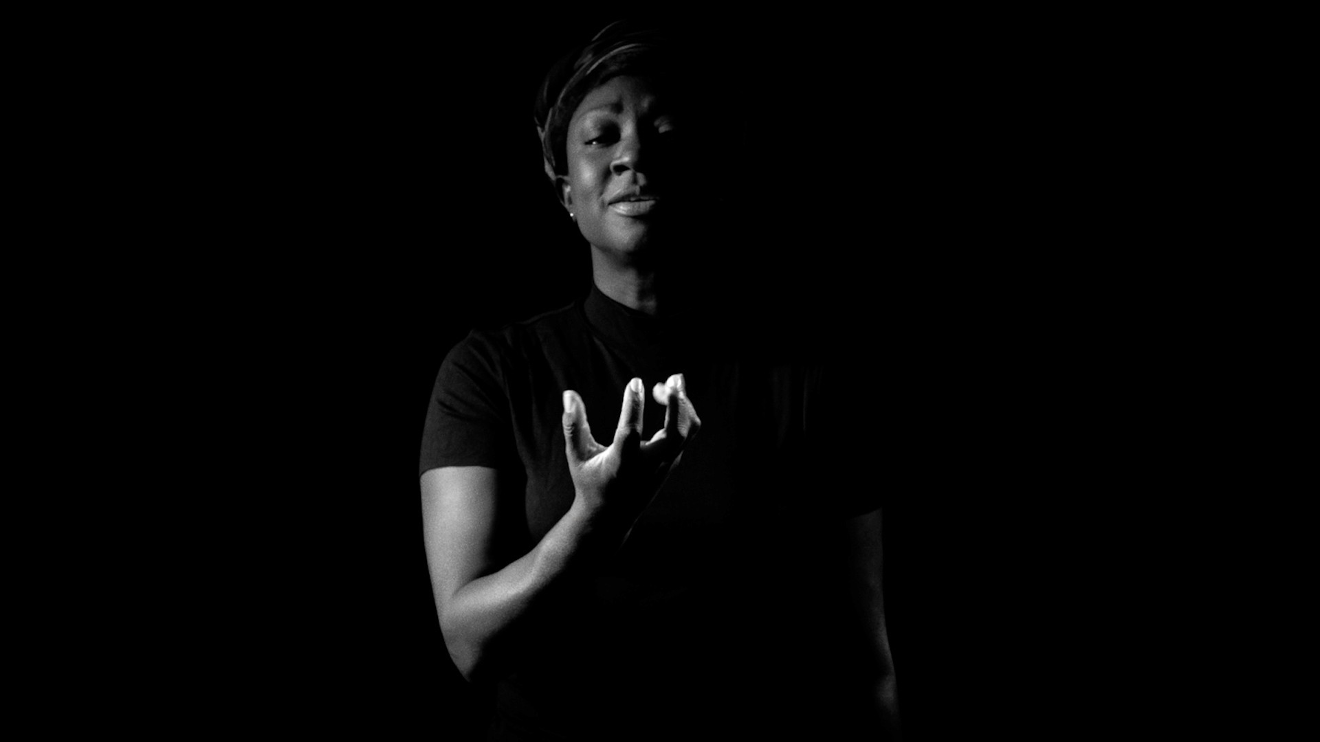 Black-and-white still photo of Natasha Ofili, a dark-skinned Black woman, with dramatic lighting on their face and hand against a dark background. She is clutching her fist softly, resembling a beating heart.