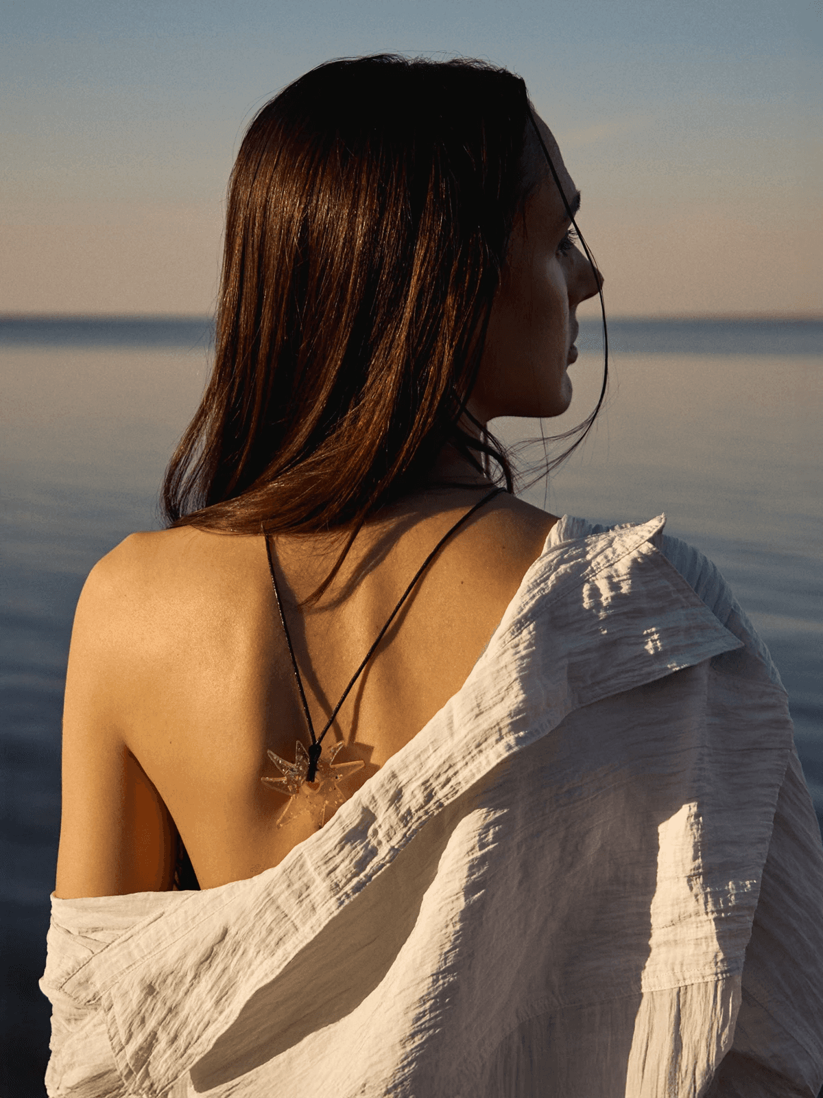 A woman with long hair gazes at a tranquil sea during sunset, wearing a lightweight white garment draped over her shoulders and a pendant necklace, creating a serene and contemplative scene by the water.