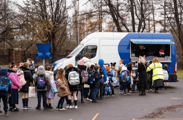 A long queue of elementary school children wearing winter coats and backpacks lining up outside a blue and white food truck
