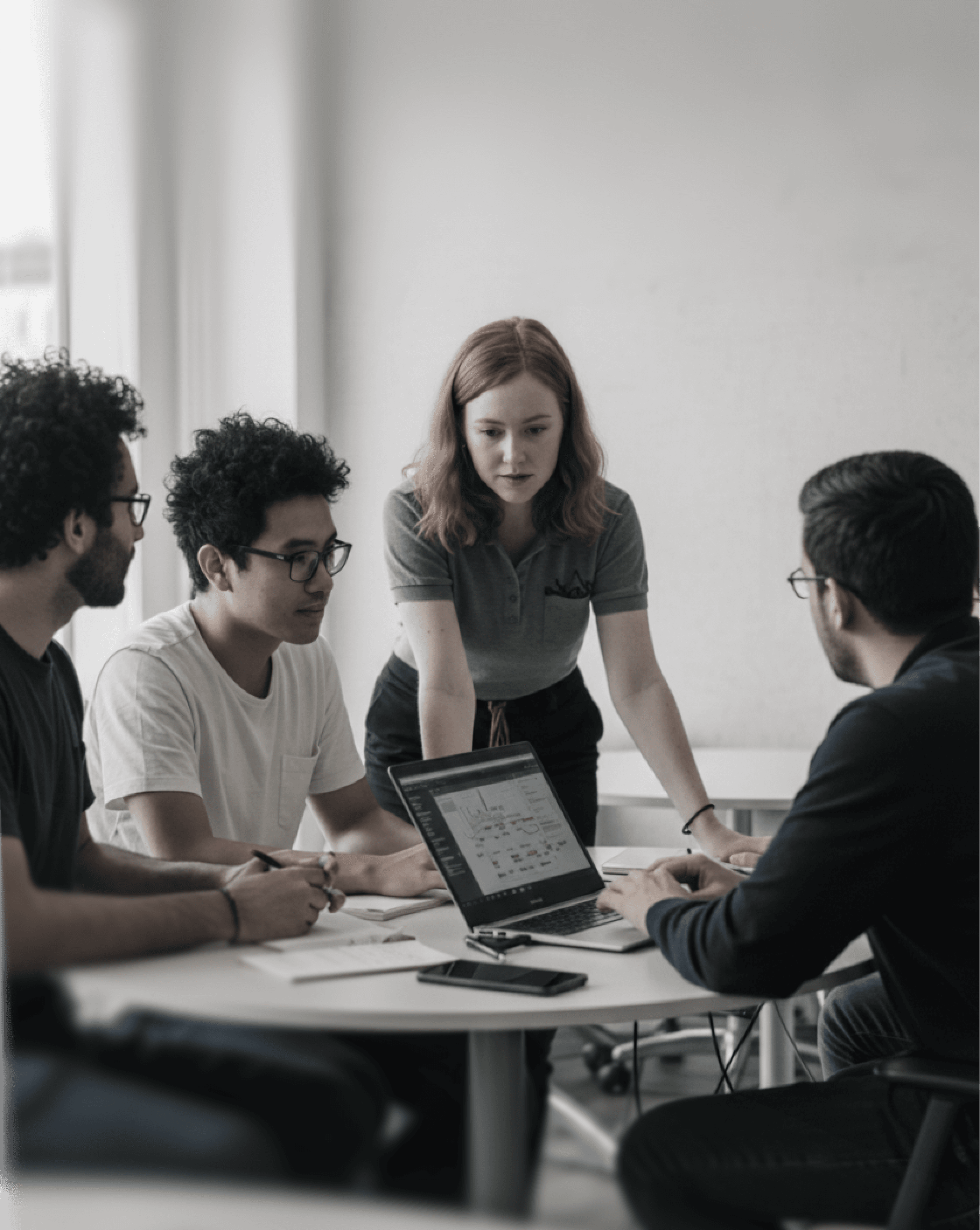 Four people focused on a laptop screen during a meeting in a bright, minimalist office. Black and white photograph.