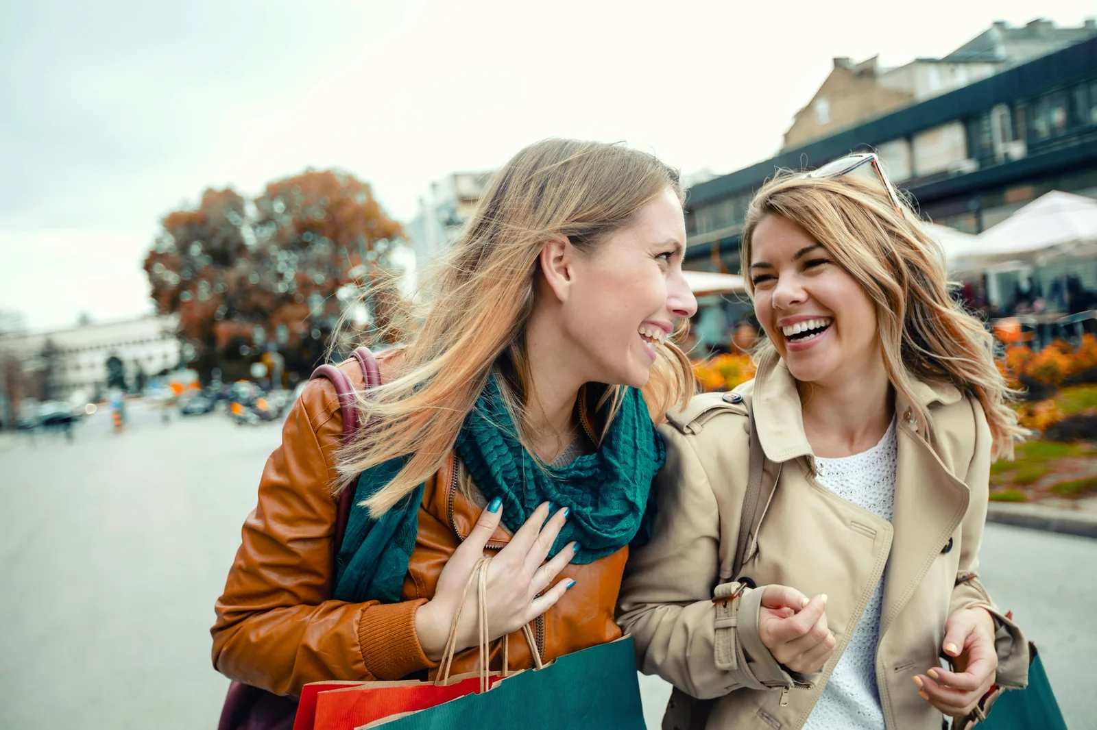 Two women shopping together in an urban waterfront setting, carrying colorful shopping bags and smiling.