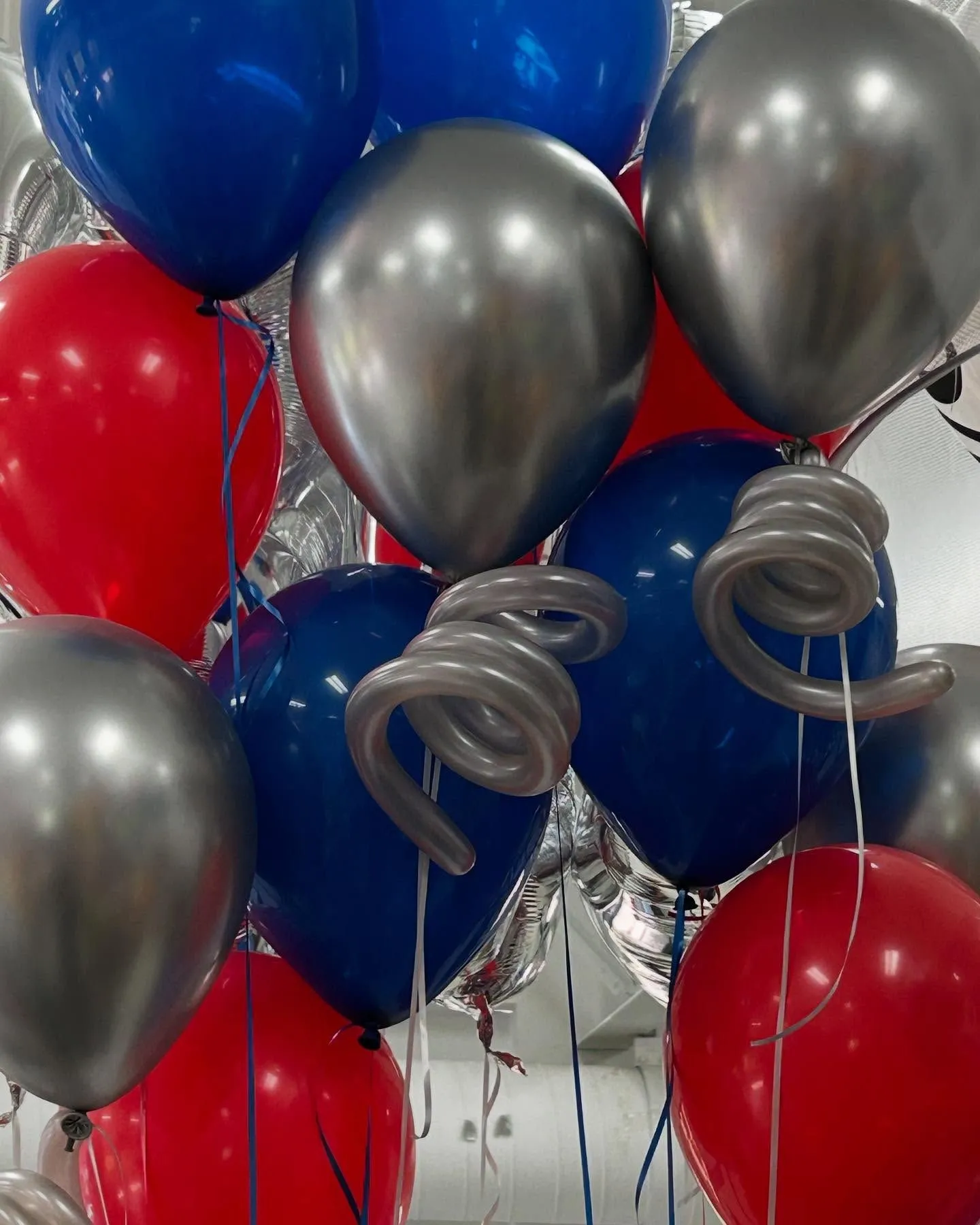 A vibrant cluster of red, blue, and silver helium balloons reflects light in a festive indoor setting, creating a lively and celebratory atmosphere.