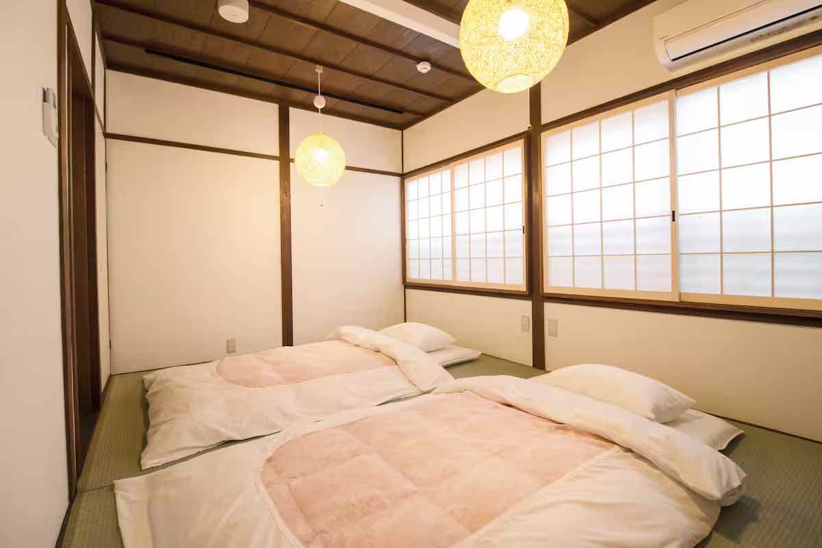 A cozy Japanese-style room features two futons on a traditional tatami floor, softly illuminated by round paper lanterns, with sliding shoji screens allowing natural light to filter in.