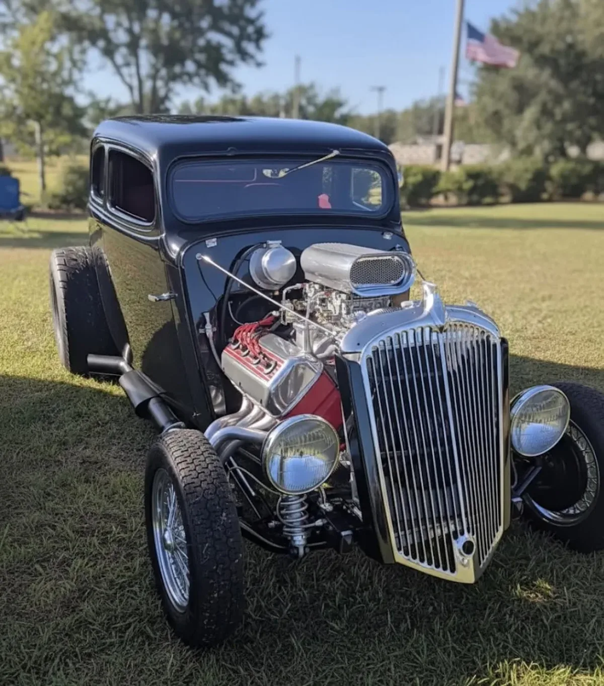 A vintage black hot rod with a polished chrome grille and exposed engine is displayed on a sunlit grassy area at the Palmetto Main Street Motor Fest in Palmetto, FL, with an American flag in the background.