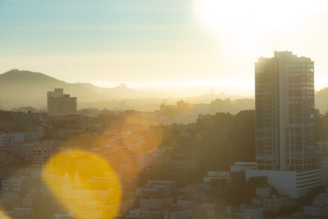 Hazy city skyline at sunrise with golden lens flare and rolling hills in the background.
