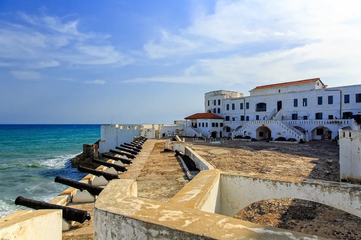 A wide shot of the historic Cape Coast Castle on a sunny day. The image features a large, paved courtyard and multi-story white buildings with red roofs. Along the stone ramparts, a long row of black cannons points out toward the bright blue sea.