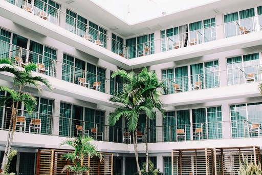Modern hotel courtyard with multiple floors of rooms, each with balconies and chairs, surrounding palm trees in a bright, tropical setting.