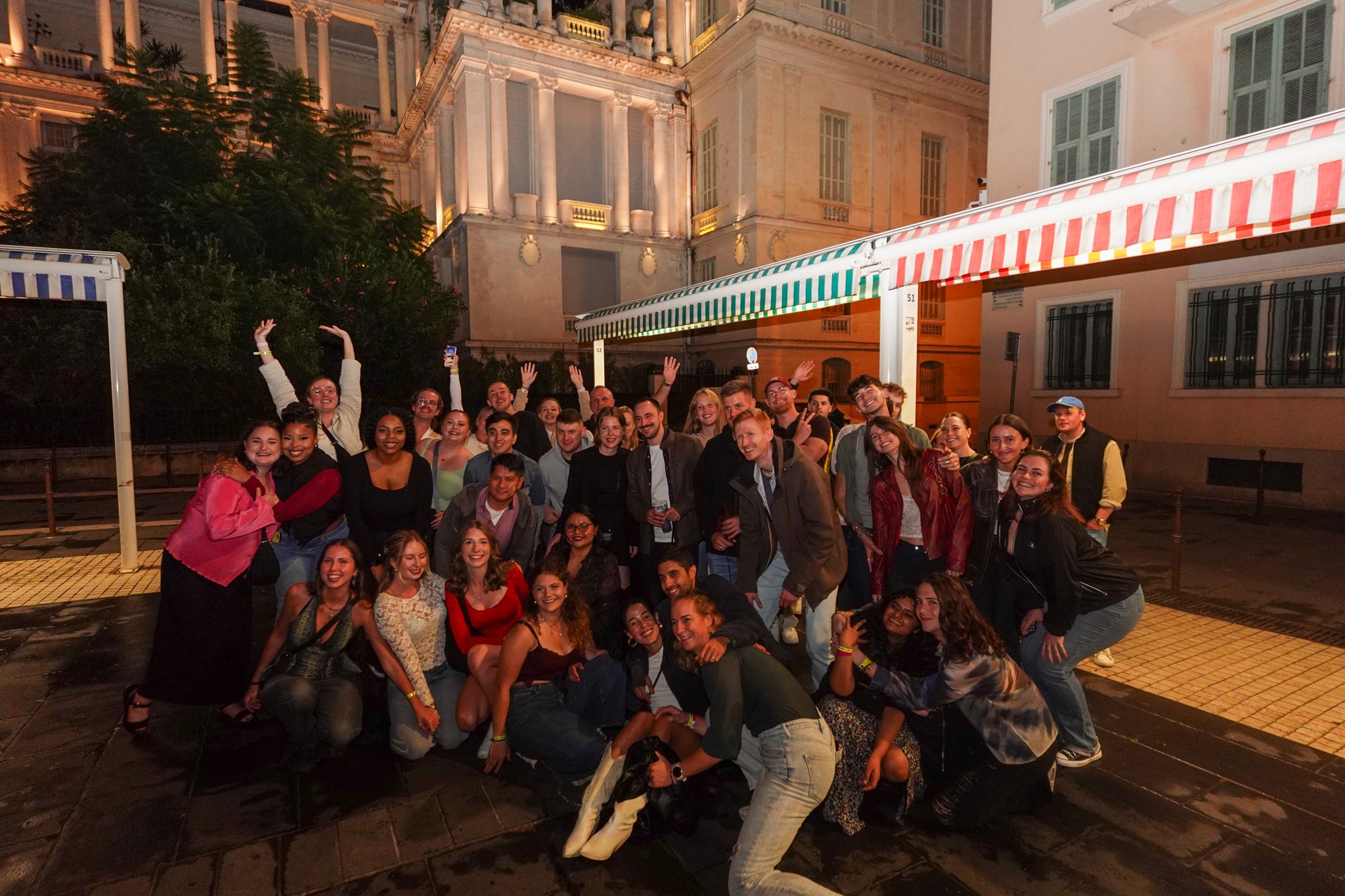 A group of international travelers walking through the streets of Nice at night, stopping at lively bars and chatting together.