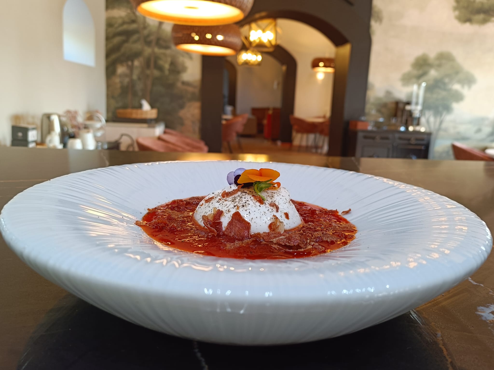 A white plate features slices of fresh mozzarella topped with black pepper and olive oil, surrounded by curls of prosciutto. The warm tones suggest a rustic dining setting.
