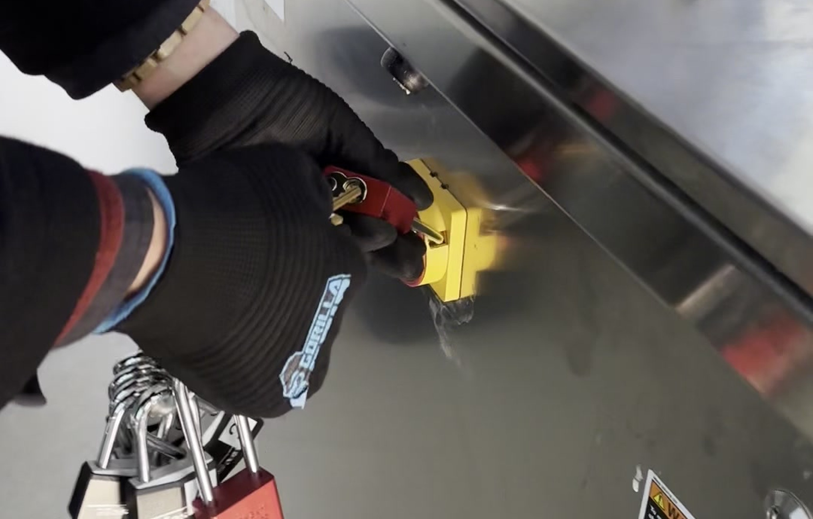 Crew member in black Gorilla Grip gloves attaching a red numbered padlock to a yellow lockout/tagout device on the stainless steel control panel of a roller coaster in Ohio, with a keyring of additional numbered padlocks visible