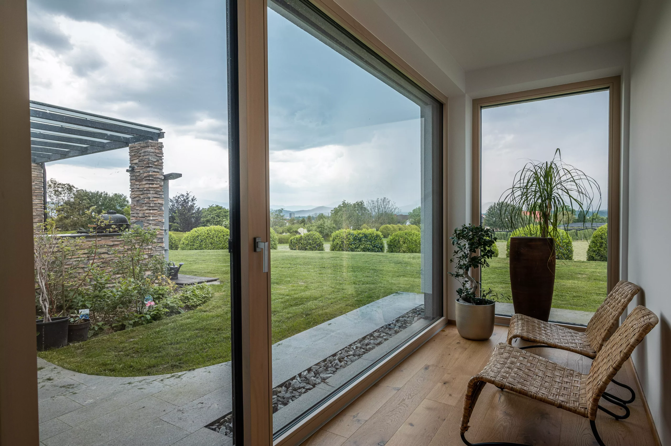 A serene outdoor seating area with two chairs, surrounded by lush greenery and a glassed-in structure.