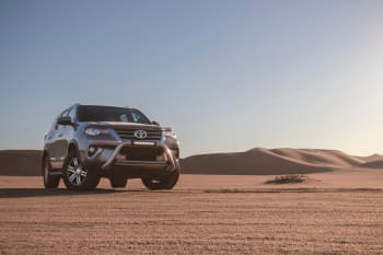 Toyota Fortuner parked on a vast desert landscape with rolling sand dunes in the background under a clear blue sky.