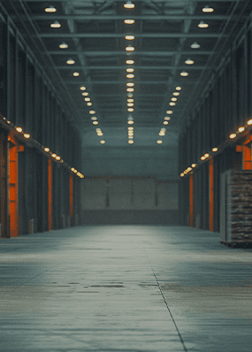 A warehouse. It has many orange parts. There are also stacks of wooden pallets in the background. Many lights are on.