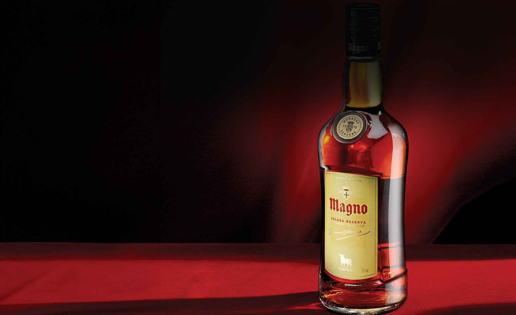 A bottle of Magno Solera Reserva brandy is displayed on a red surface against a dark background. The lighting creates a warm, elegant ambiance.