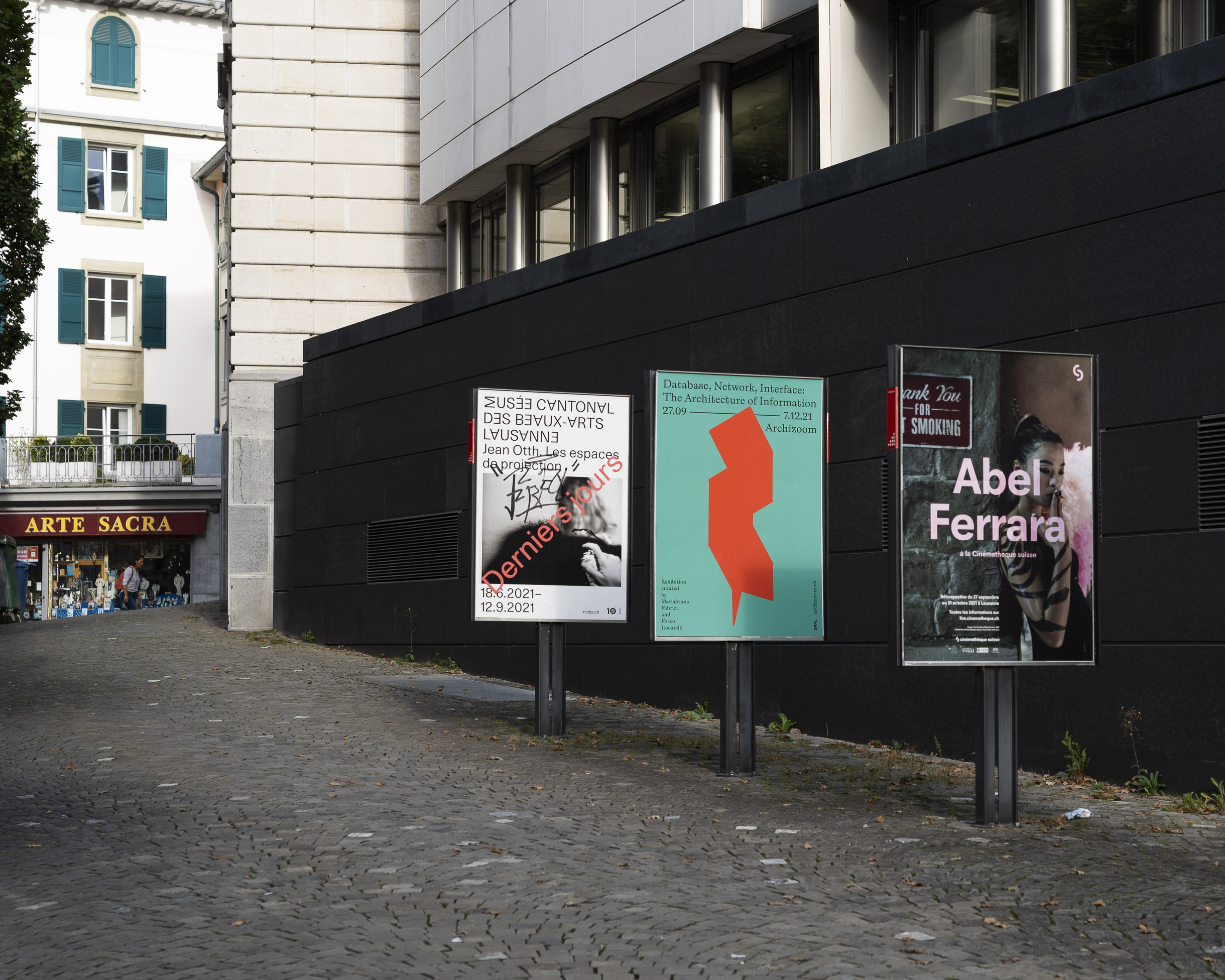 Photo en ville de Lausanne montrant 3 affiches dans une rue passante dont celle du milieu montre l'affiche de l'exposition Archizoom EPFL