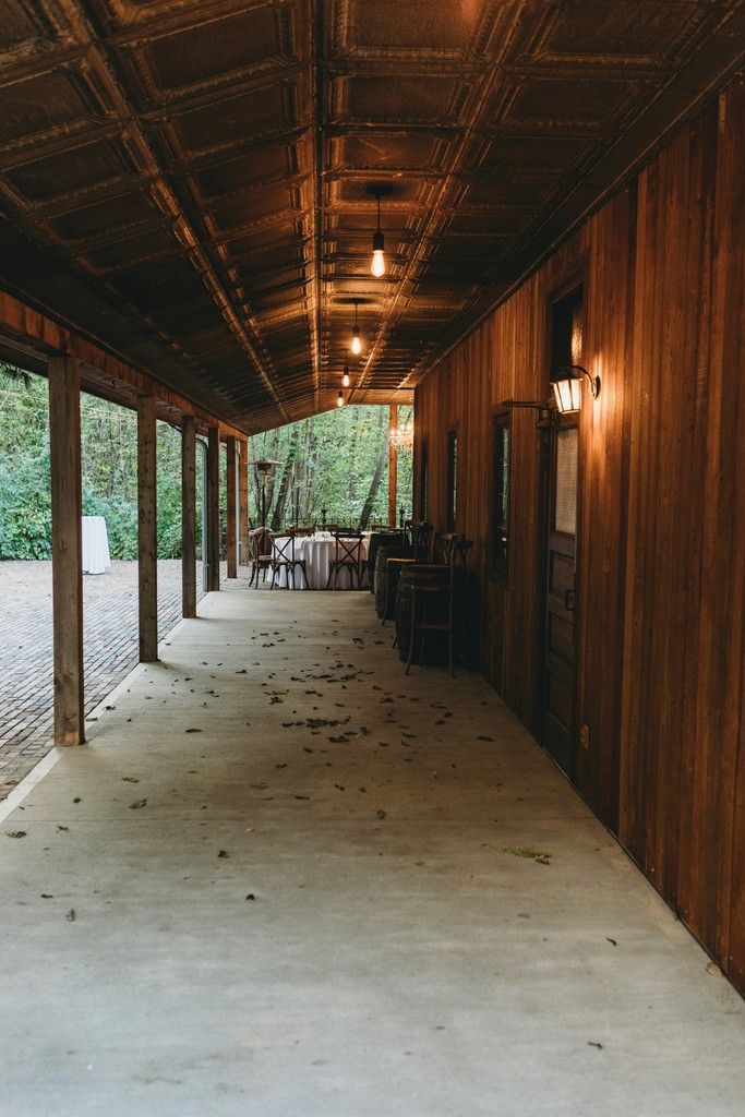 Covered wooden porch of a rustic wedding venue with pendant lights, tin ceiling panels, and a wooded backdrop leading to the reception area