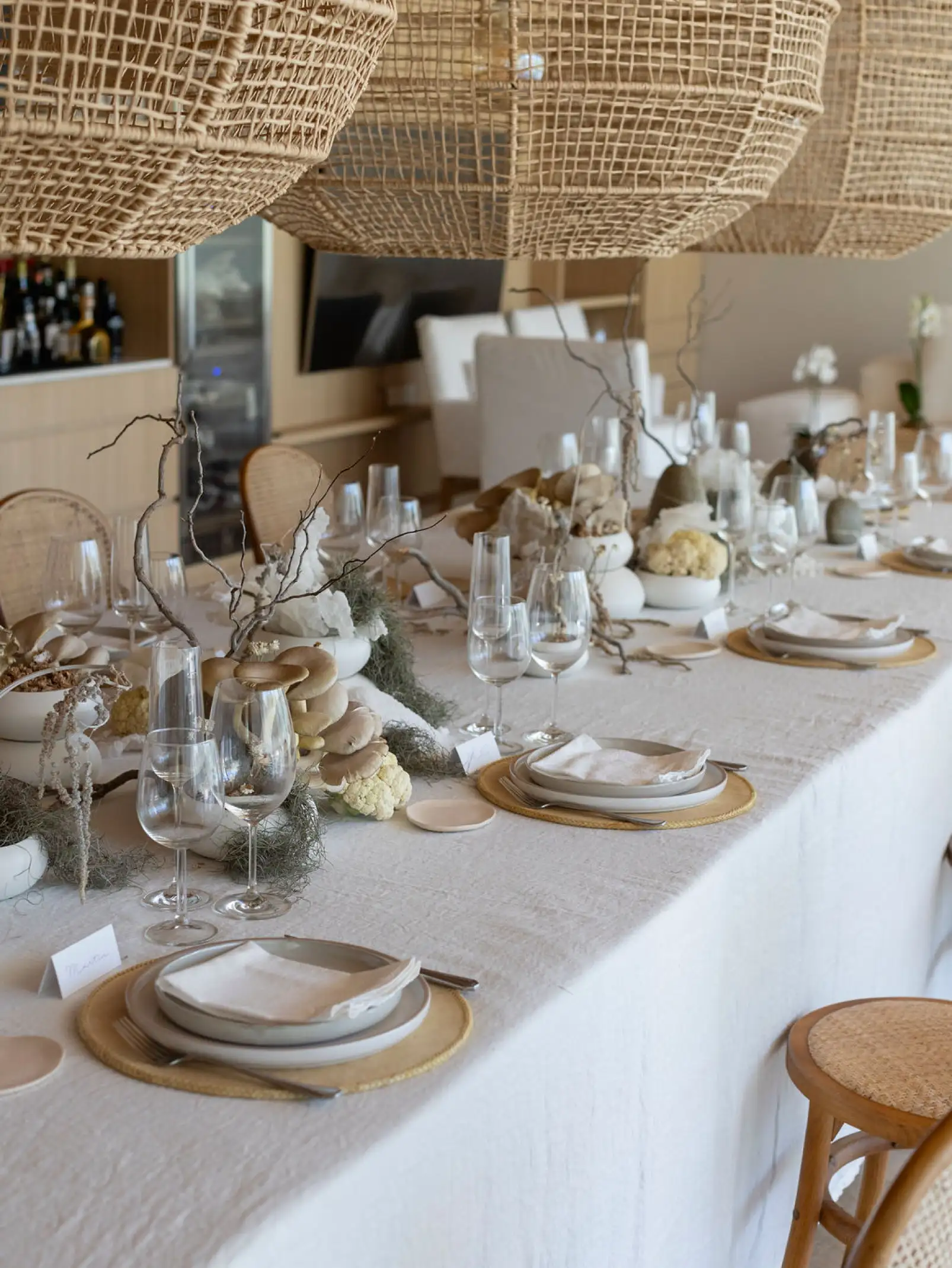 Vista de mesa larga decorada con hongos, ramas secas y elementos naturales en tonos neutros, con vajilla minimalista y lámparas de mimbre colgantes.