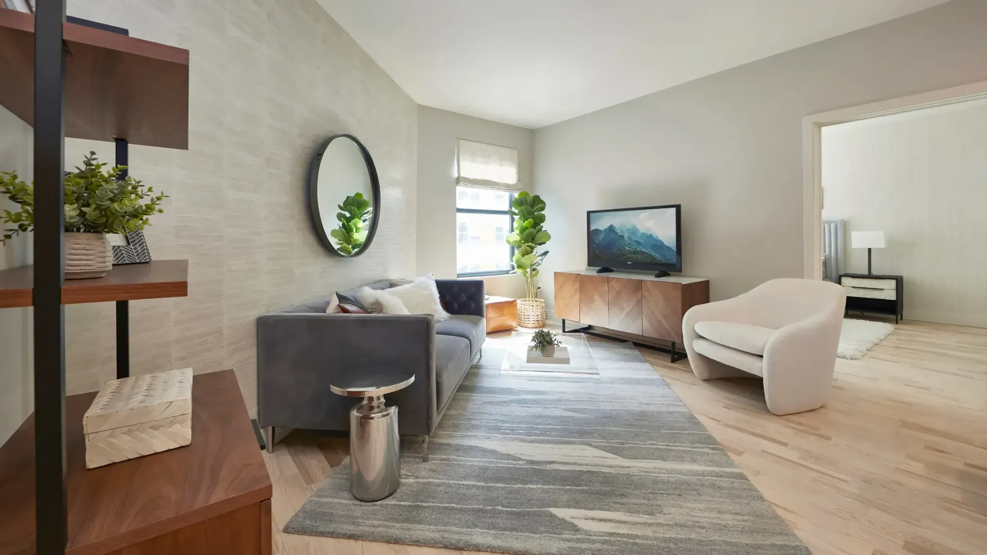 A stylish living room featuring a gray sofa, a round mirror, a TV, and modern decor, with natural light coming in.
