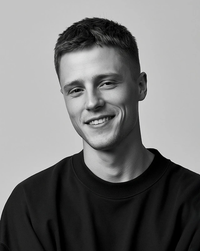 A professional, black-and-white studio portrait of a smiling young man with short, styled hair, wearing a black shirt.