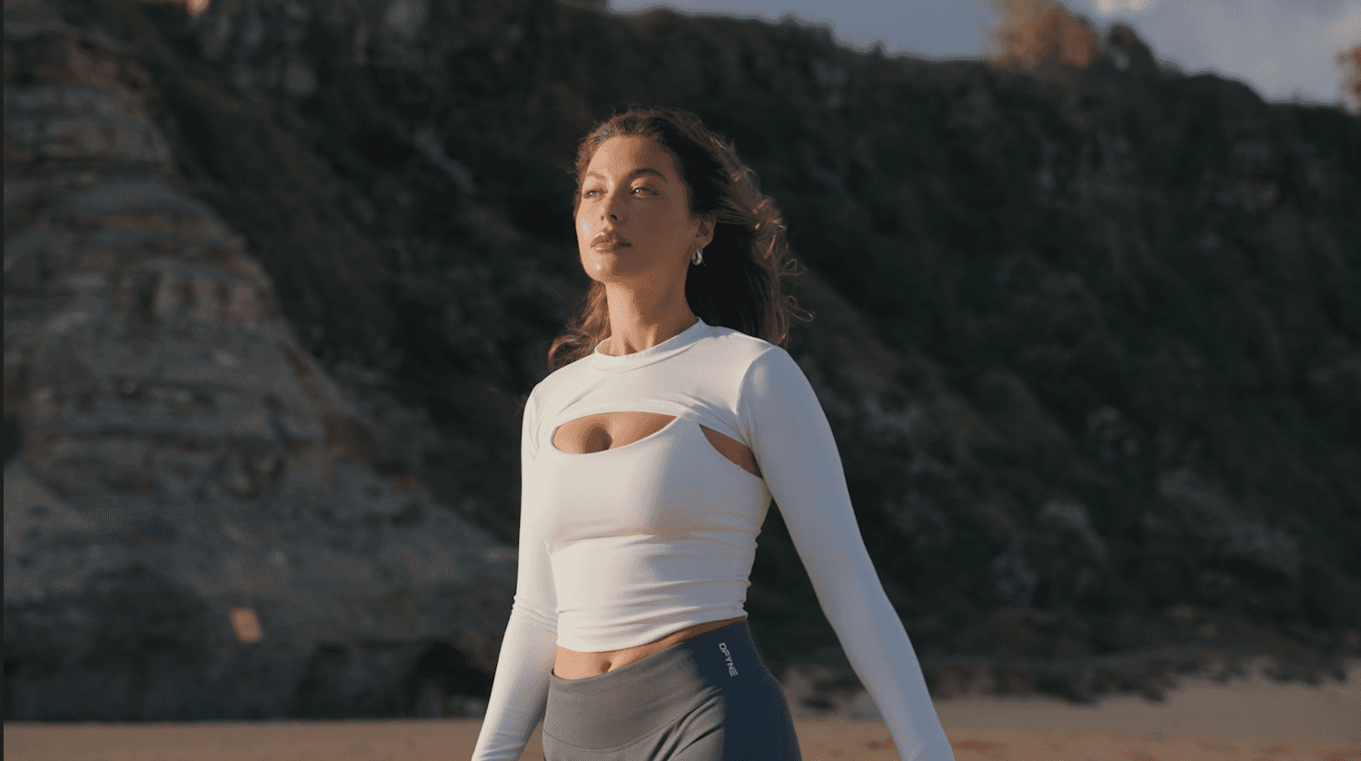 Woman in white cutout top and dark pants by cliffs.