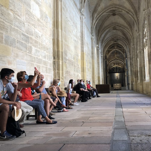 People sitting on benches inside a stone-arched corridor, some are wearing face masks and taking photos.
