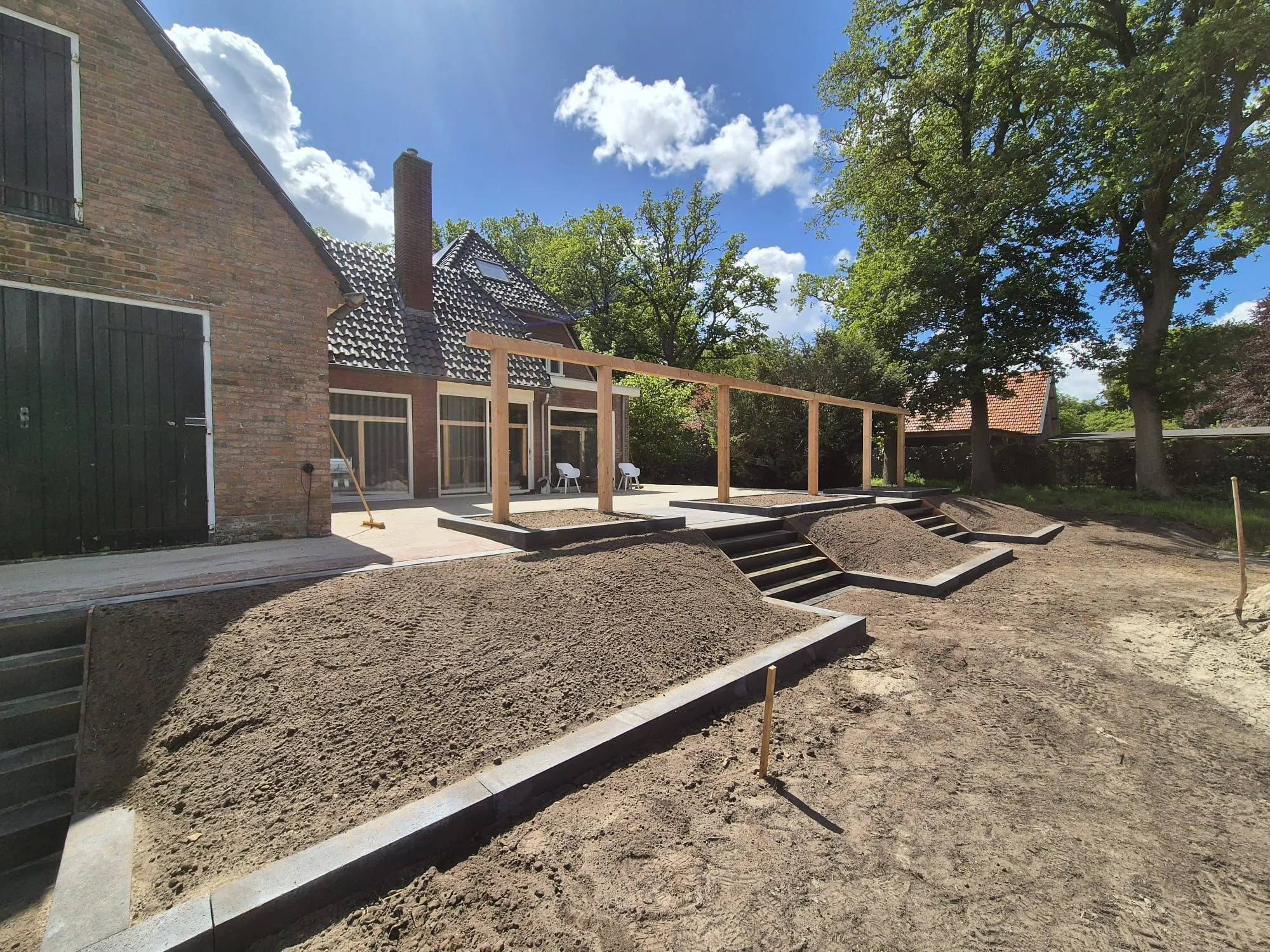 Rustiek verhoogd terras met trap, houten pergola en drie verhoogde tuinbedden in aanleg bij bakstenen huis onder blauwe lucht.