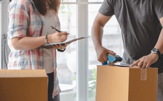 A man and woman, dressed as office movers, are carrying boxes into a house, by Extra Space