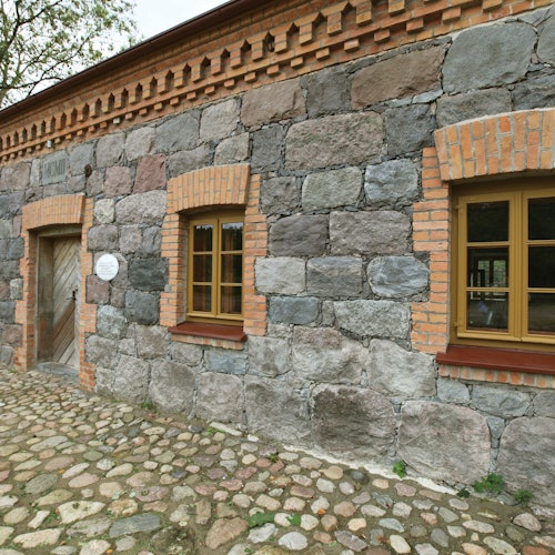 A stone building with a wooden door and two windows framed by brick, situated on a cobblestone pathway.