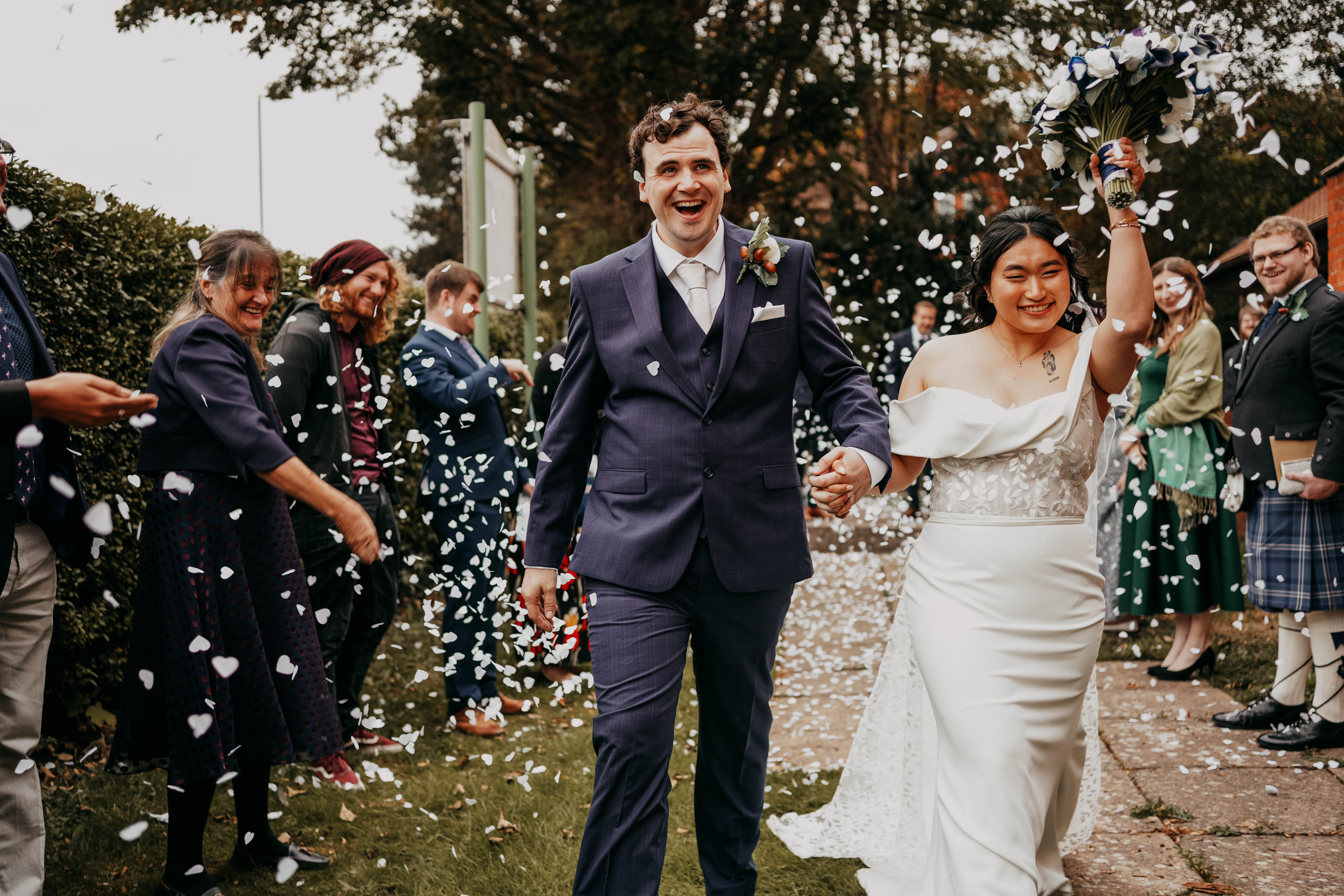 The newly married couple walks hand-in-hand outdoors as guests shower them with confetti. The groom is laughing exuberantly, and the bride is smiling as she raises her bouquet in the air in celebration.