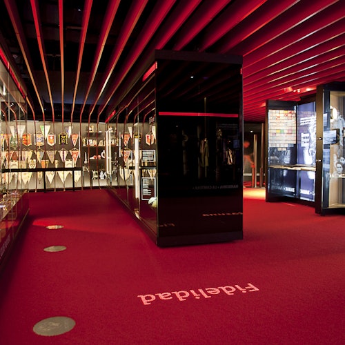 A museum exhibit with red carpet, black display cases, and various artifacts. The ceiling has red lighting and parallel beams.