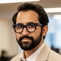 Joaquin, a Latino with dark mustache and beard wearing glasses and a white shirt and beige blazer, smiling at the camera.
