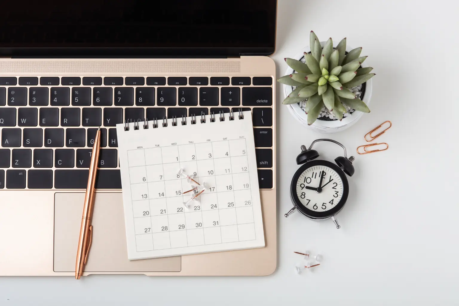 Scheduling automation workspace featuring a laptop, desk calendar, clock, succulent, and copper pen on a clean desk.