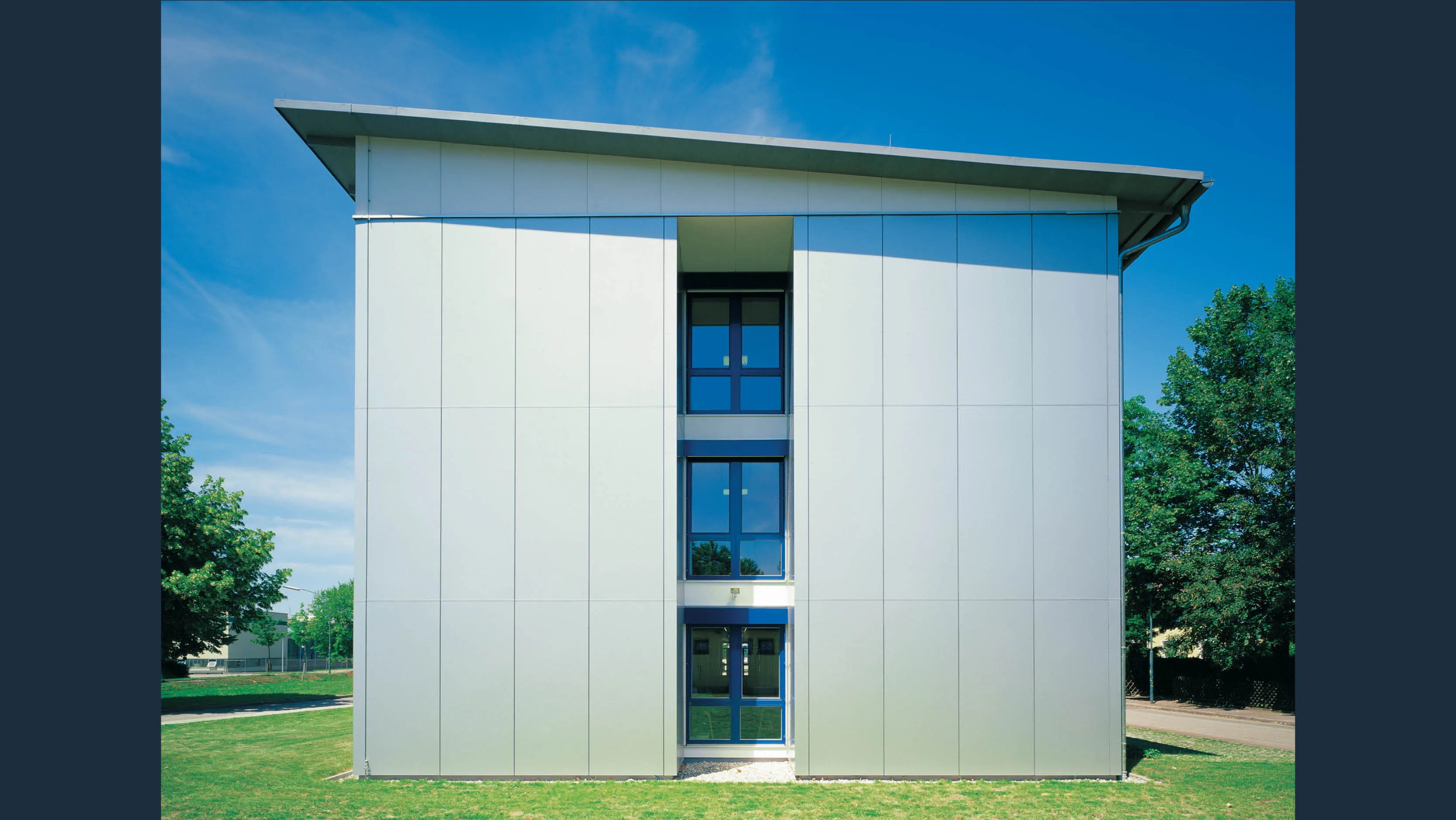 Modernes, helles Gebäude mit Aluminiumfassade, drei vertikal angeordneten Fenstern und einem schrägen Dach im Sommer.