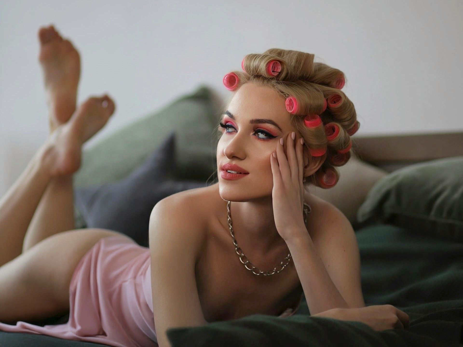 A blonde woman with pink hair rollers and pink eyeshadow lying on a bed with her feet up, wearing a pink silk garment and a silver chain necklace.