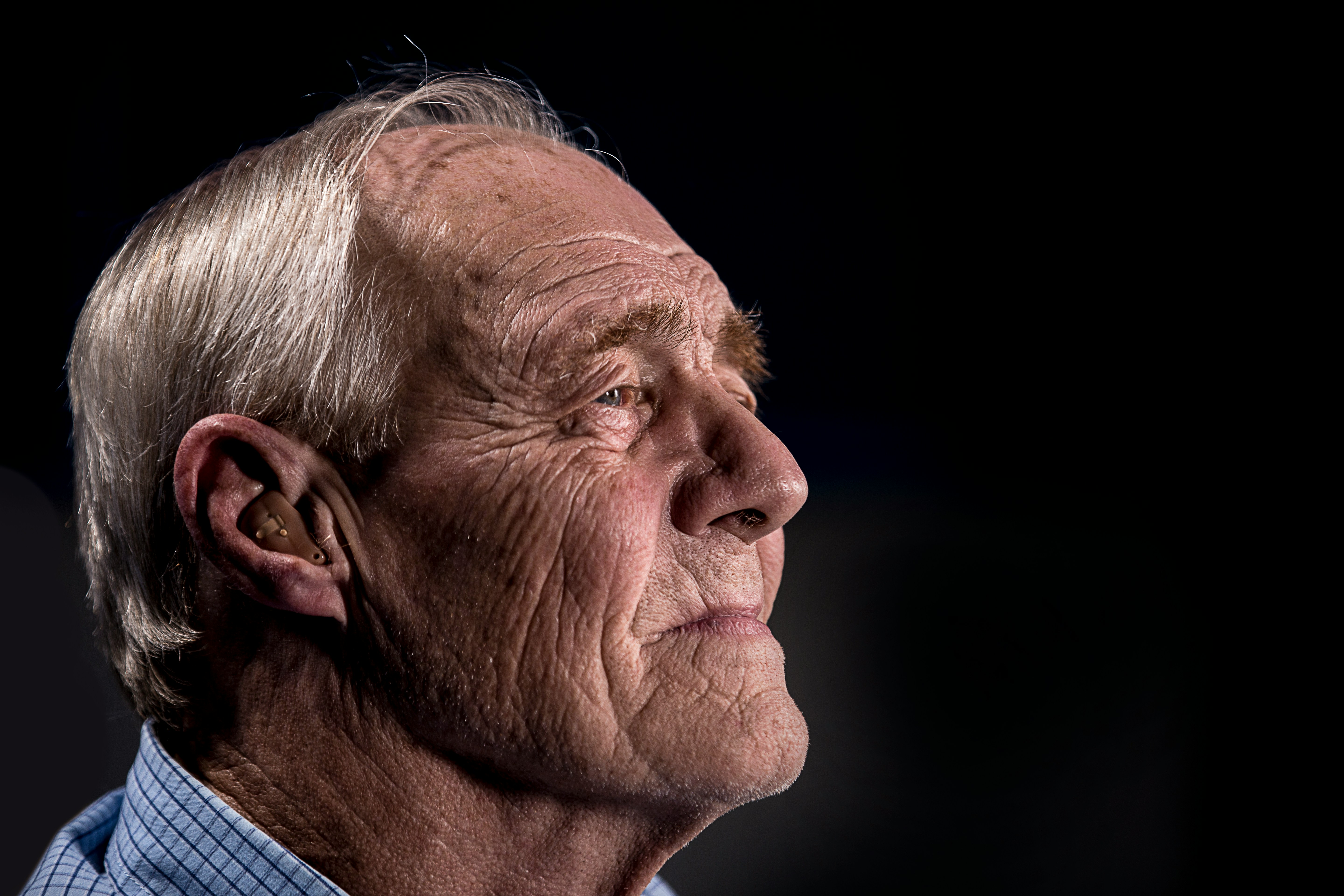 Side profile of an elderly man with a thoughtful expression, wearing a hearing aid. His face and features are illuminated against a dark background, highlighting wrinkles and the texture of his skin, conveying a sense of wisdom and experience.