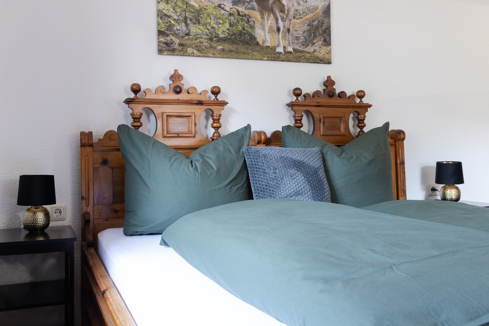 Ein gemütliches Schlafzimmer verfügt über ein Doppelbett aus Holz mit kunstvoll geschnitzten Kopfteilen und blaugrüner Bettwäsche, flankiert von schwarzen Nachttischen mit kleinen Lampen, unter einem Wandbild mit Naturmotiven, das einen Hirsch darstellt.
