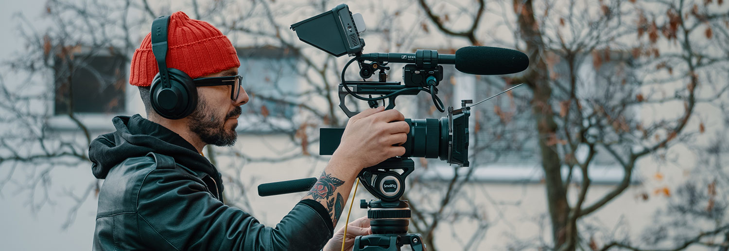 A camera man in red beanie and leather jacket looks at the camera display