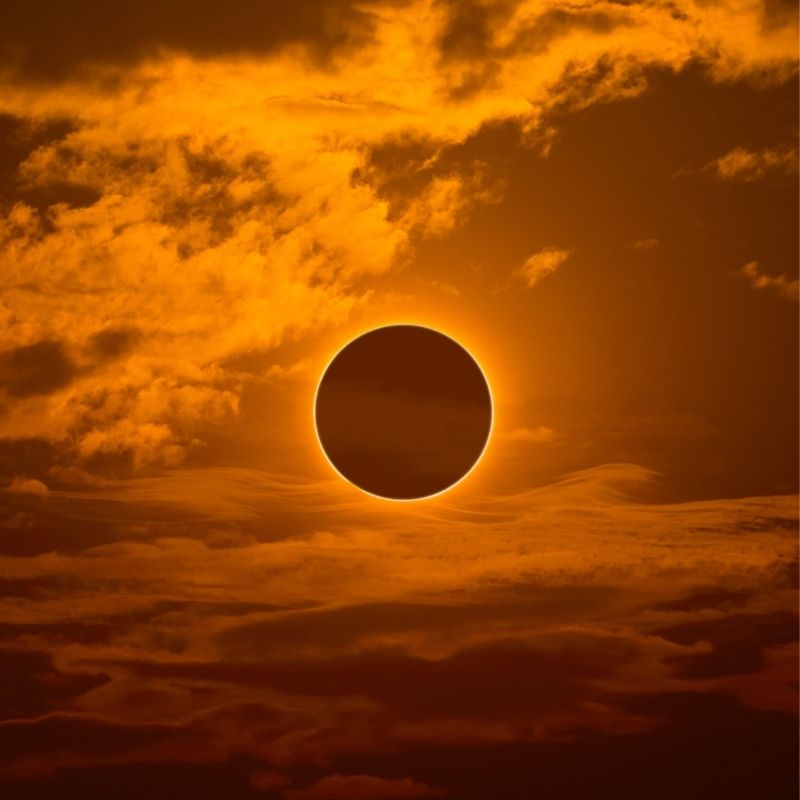A total solar eclipse with the Moon fully obscuring the Sun, surrounded by a bright golden corona against dramatic orange clouds in the sky.