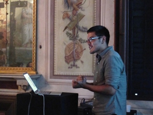 A photo of a person giving a lecture while sitting in a room with frescoes on the walls.