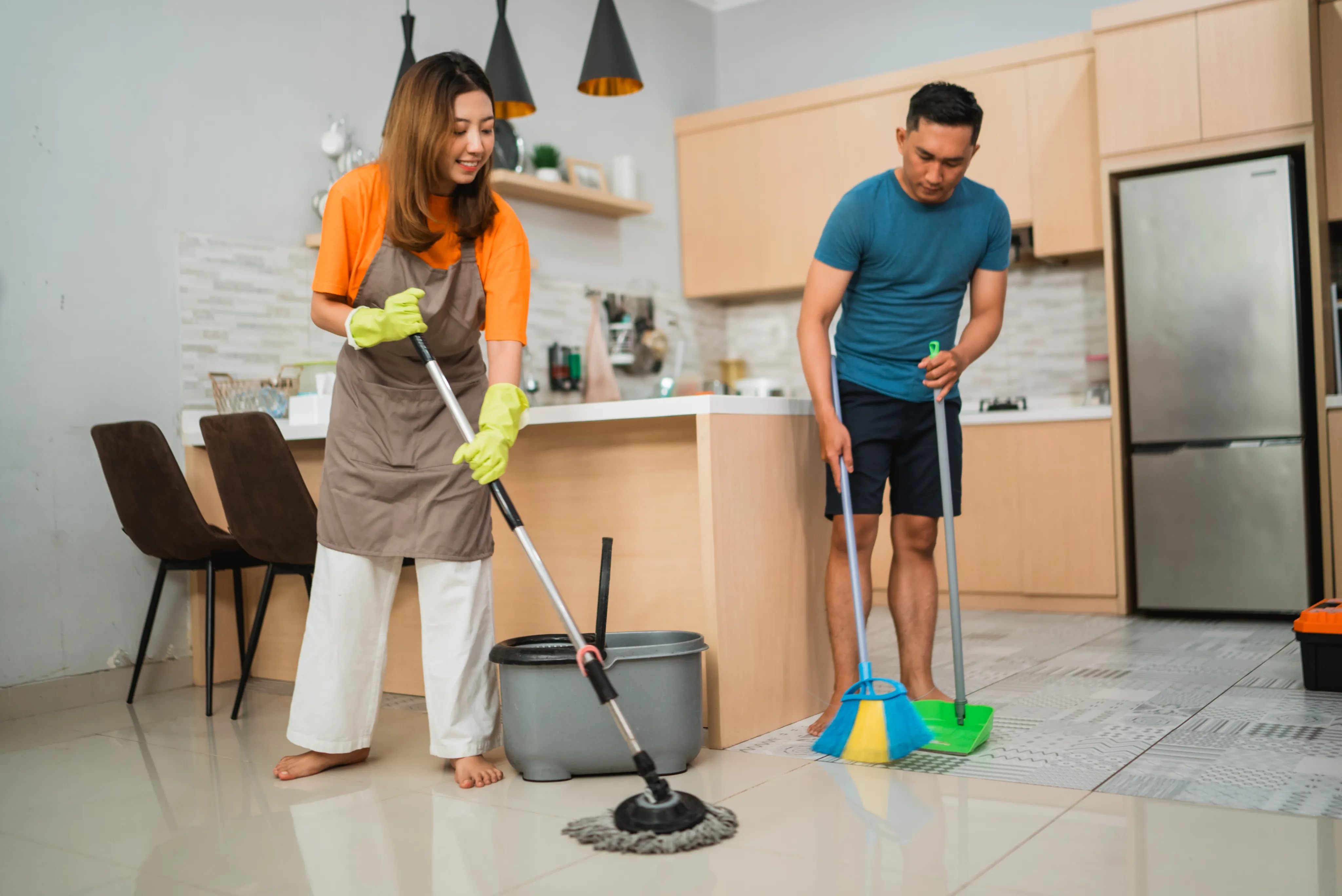 A young couple working together to clean their home kitchen using a mop, broom, and bucket.