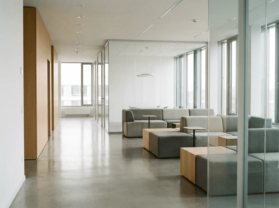 Heller, moderner Loungebereich in einem Büro mit grauen Polstersofas, Beistelltischen aus Holz, einem hochglänzenden Estrichboden und großen Glaswänden im Hintergrund.