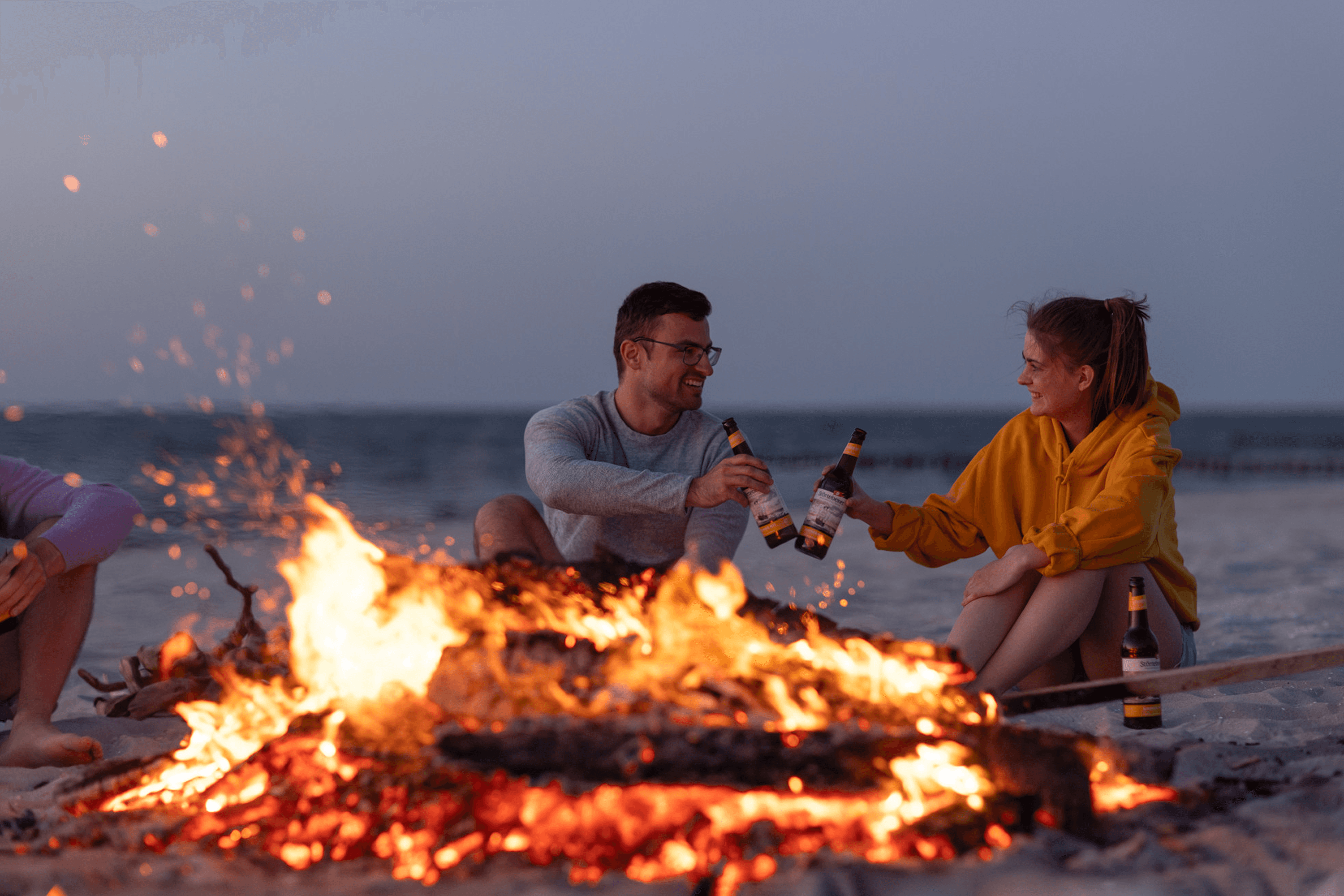 Couple toasting bottles by a beach bonfire