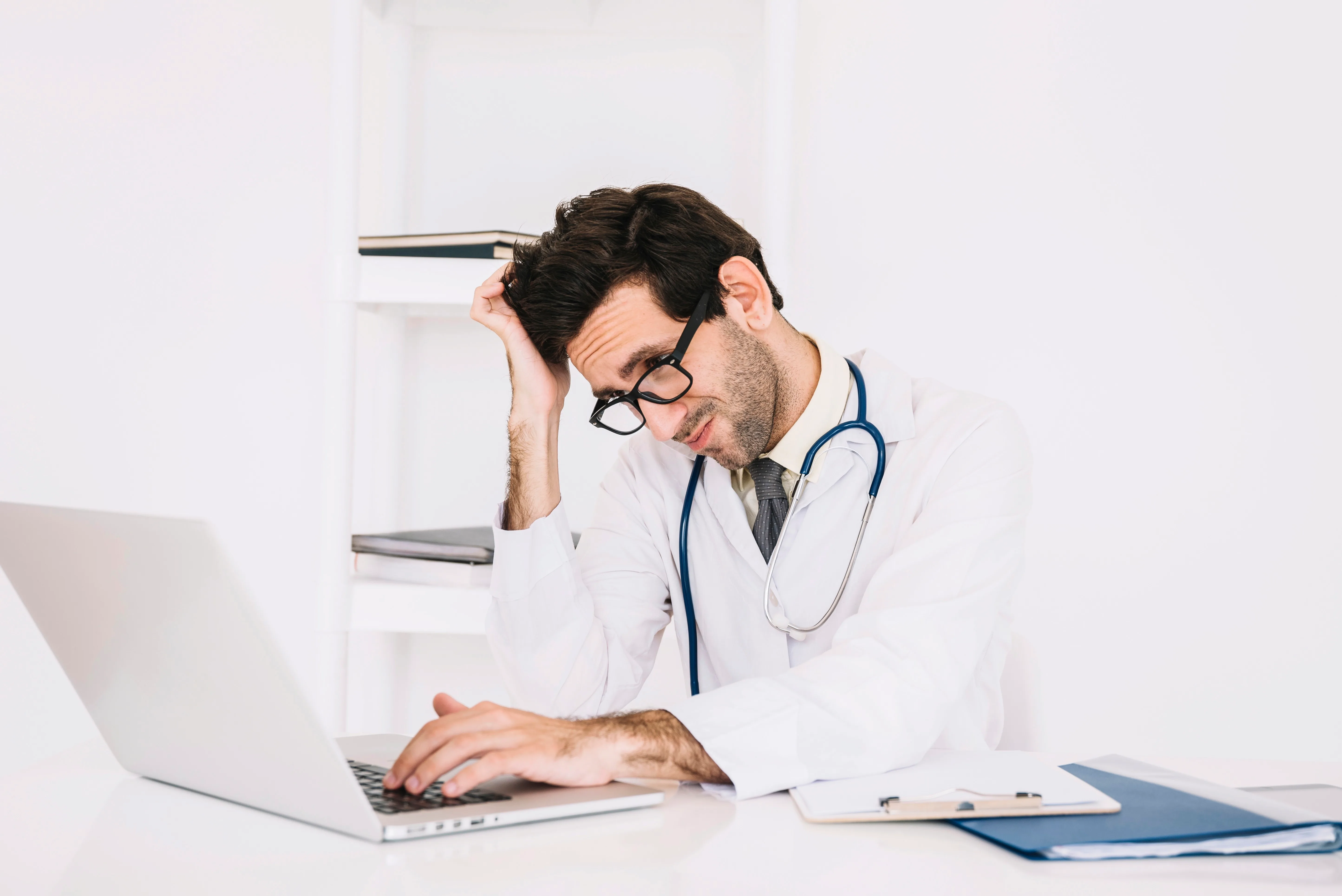 A male doctor in a white coat and stethoscope appears stressed while working at a laptop, holding a pencil to his head with papers and a clipboard on the desk beside him.