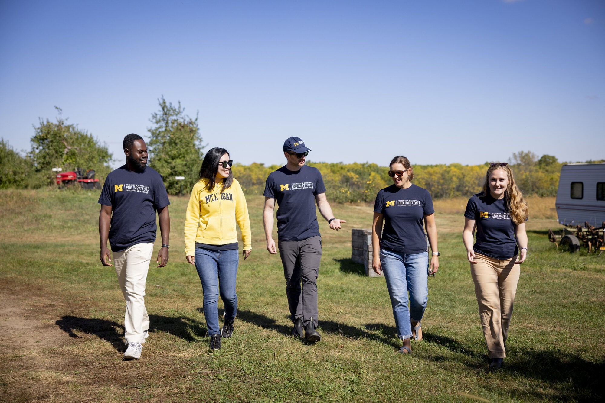 A group of five people walking together outdoors in casual attire, with a clear blue sky in the background.