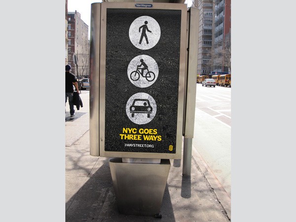A metallic sign with three circular icons depicting walking, cycling, and driving is mounted on a sidewalk. The sign reads “NYC GOES THREE WAYS” below “3WAYSTREET.ORG”. A person walks past the sign on the left side of the image. Buildings and vehicles are visible in the background.